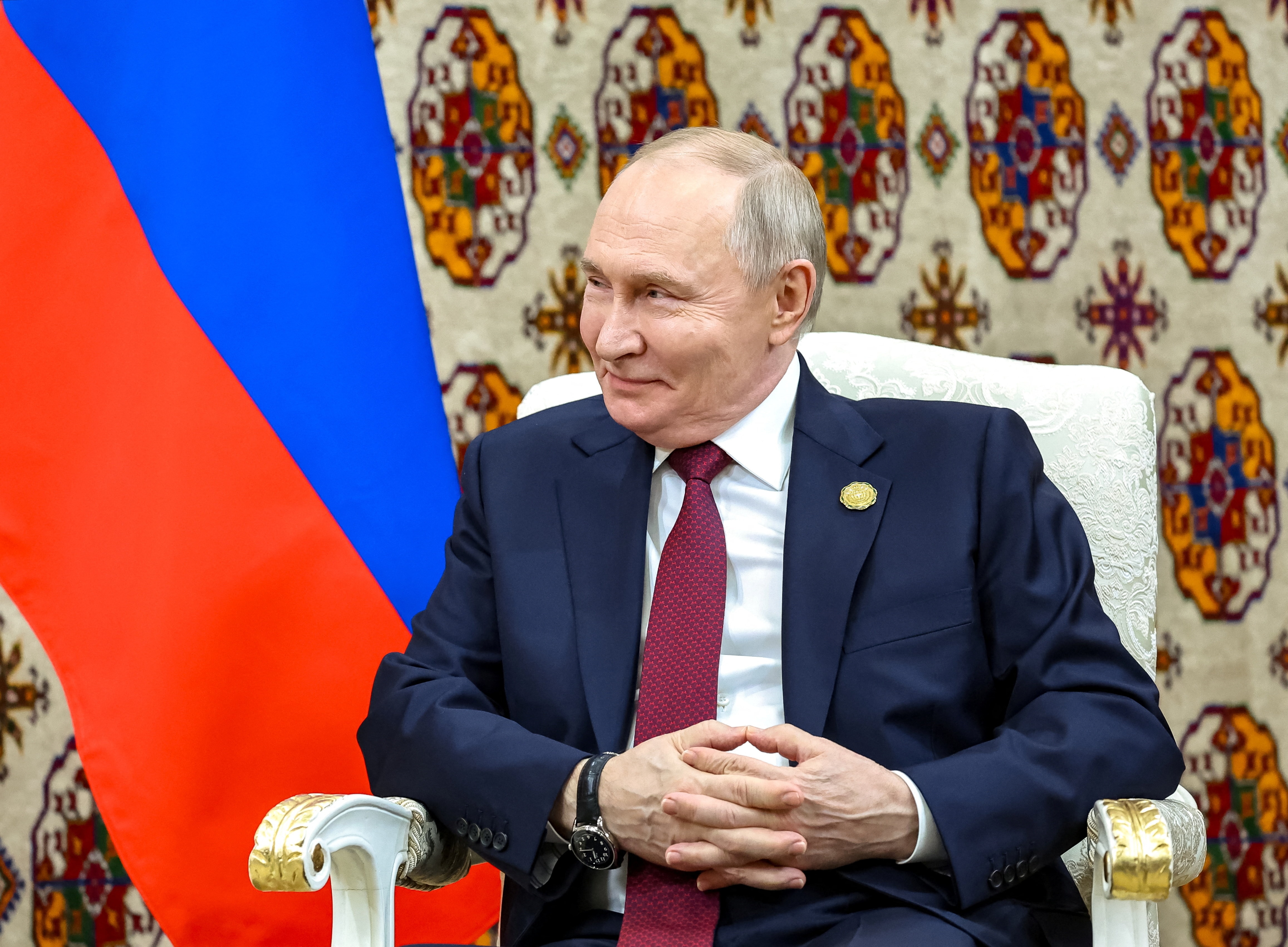 Vladimir Putin sentado em uma cadeira com uma bandeira russa atrás dele.