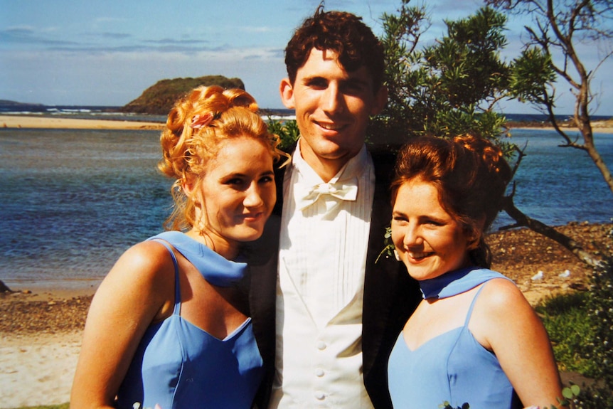 A man in a tuxedo stands between two women in matching blue dresses on a beach