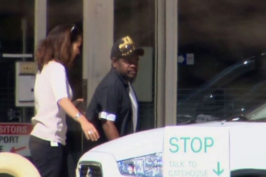 An Aboriginal man walks out of a building towards a white car with a woman behind him.