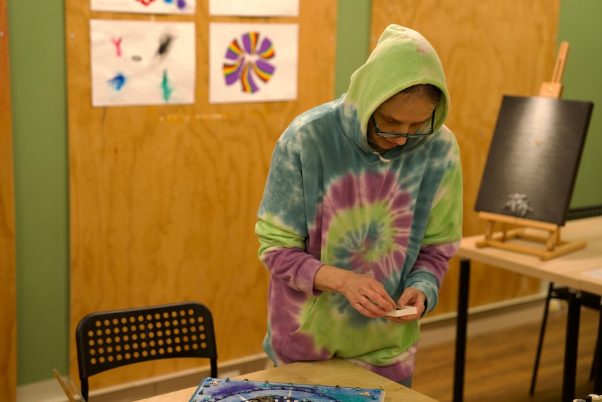 A middle aged white woman with black glasses wearing a rainbow hoodie. She is standing at a table making art