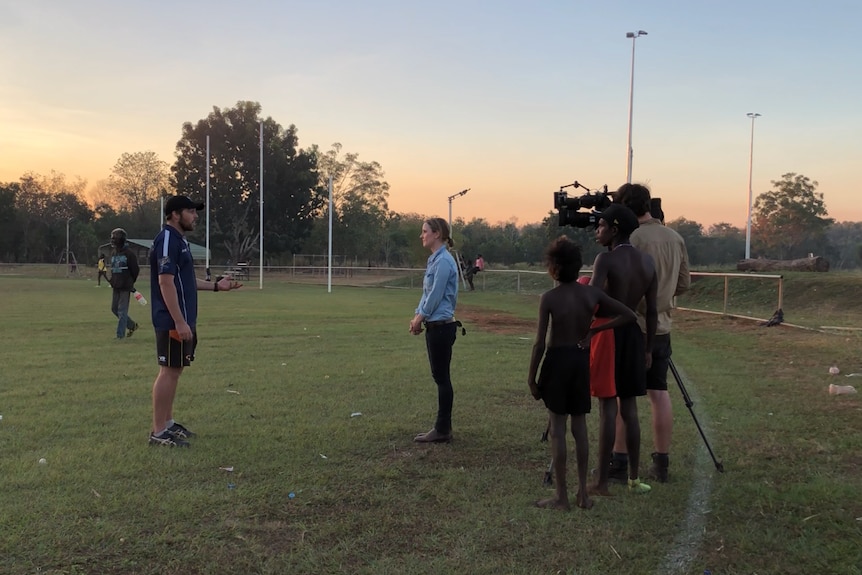 A man is interviewed for a television story on a football oval at dusk.