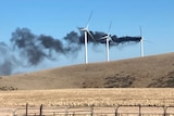A series of wind turbines among farmland with black smoke coming from one of them