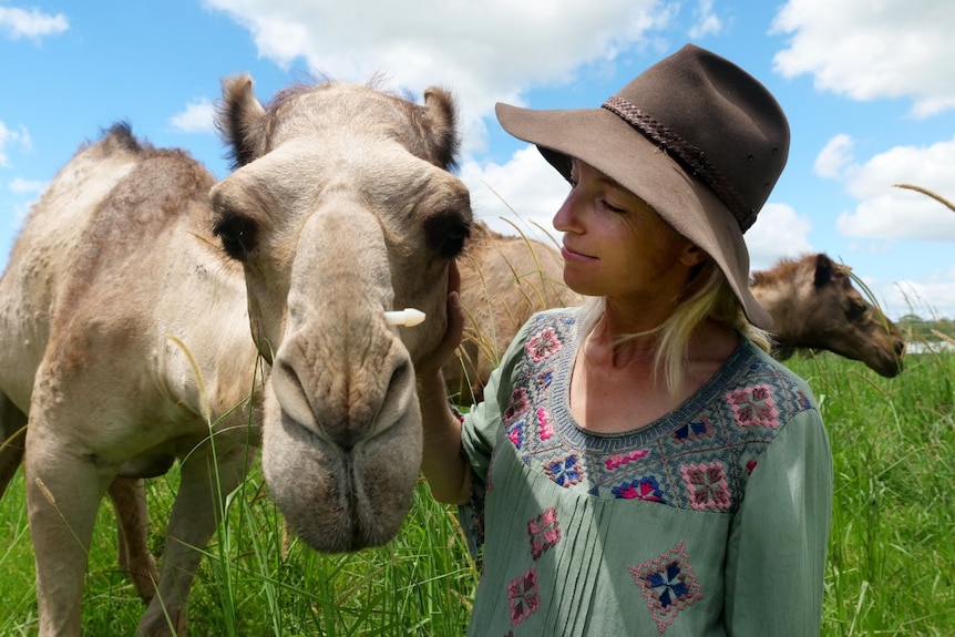 A blonde woman in a hat next to a camel with her hand on its head