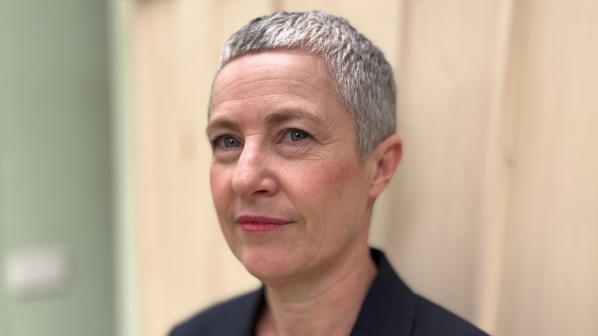 Kate Colvin, with short grey hair and wearing a black blazer, stands against a beige wall and looks at the camera.