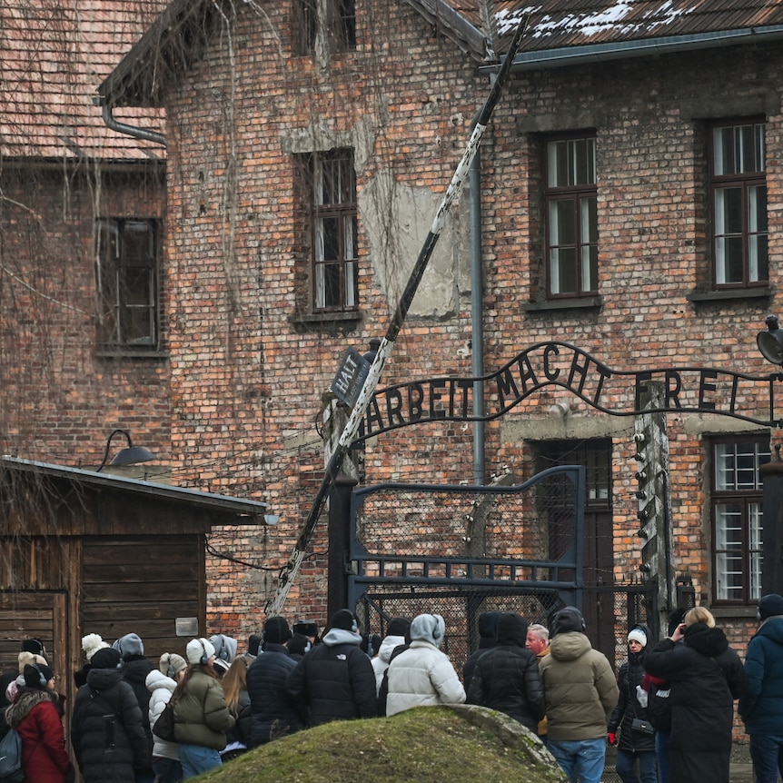 Small crowd wearing jackets in front of 'Arbeit Macht Frei' sign at the main entrance gate of Auschwitz
