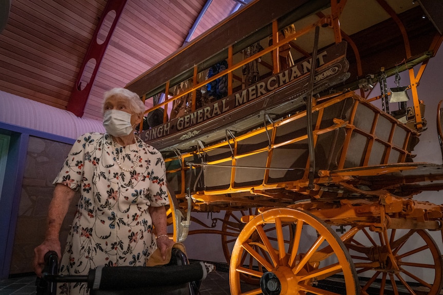 An elderly woman in a mask stands in front of an old wooden buggy in a museum.