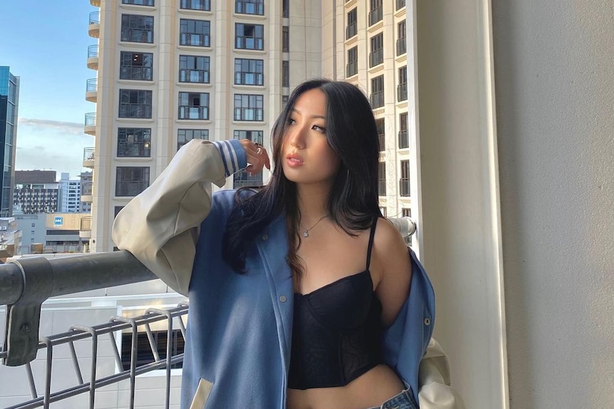 Cherry Park standing against a balcony fence. She's looking off to the side and wearing a blue and white jacket, black singlet 
