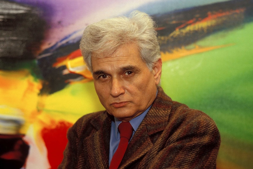 A man with grey hair, wearing a brown suit, blue shirt and red tie, stands in front of a brightly coloured painting.