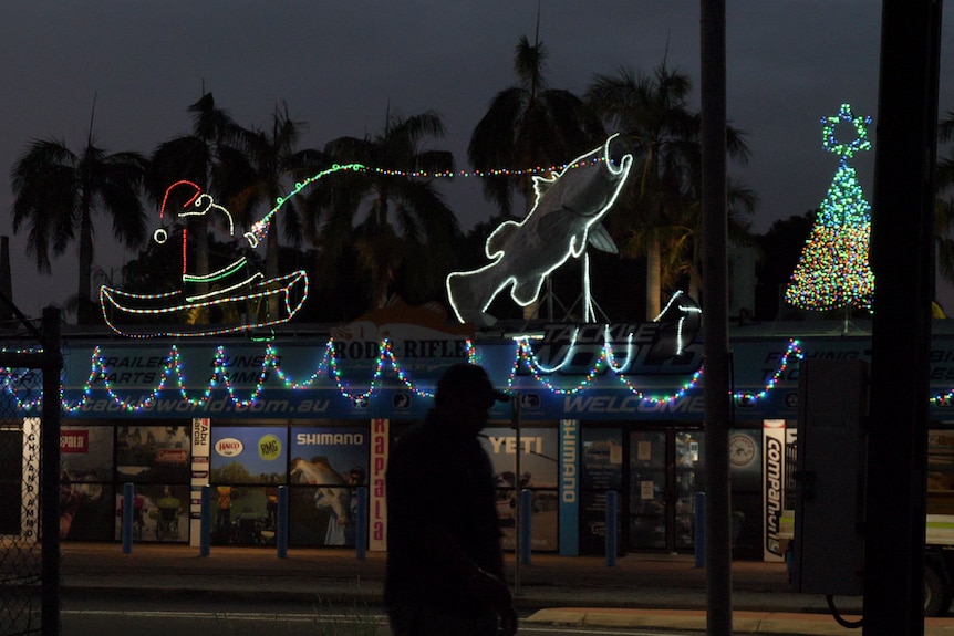 In a Christmas lights display on top of a fishing tackle shop at night, an illuminated Santa is pulled along by a barramundi. 