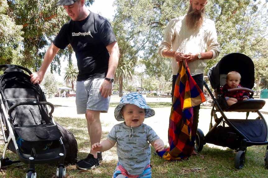 Two fathers stand in a park one toddler stands and another baby sits in a pram for a dads' support group.