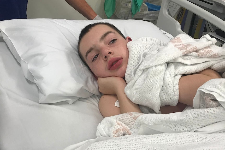 A boy with brown hair lying in a hospital bed, wrapped in a white hospital blanket, neutral expression.