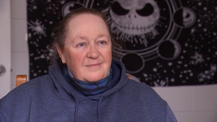 A woman wearing a blue jumper looks off camera. Behind her is a black and white poster