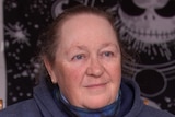 A woman wearing a blue jumper looks off camera. Behind her is a black and white poster