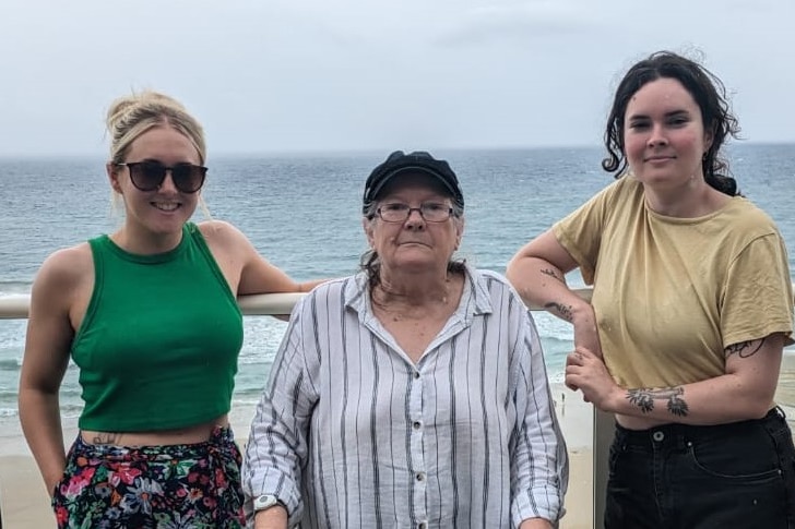 Three women stand together on the sidewalk with the ocean in the background.