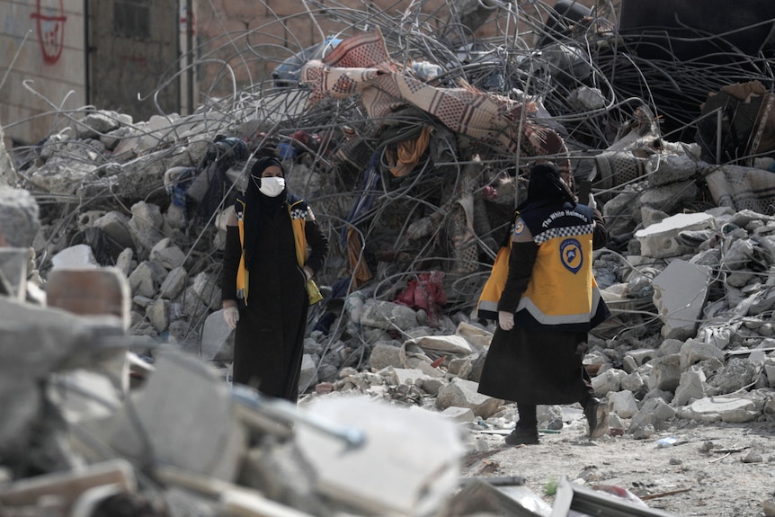 Woman volunteers in white helmets walk at the site of a damaged building.
