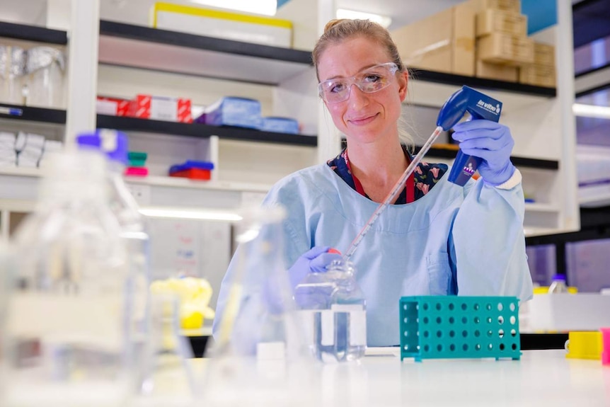 Kirsty Short, wearing a lab coat, blue gloves and goggles, holds equipment in a lab.