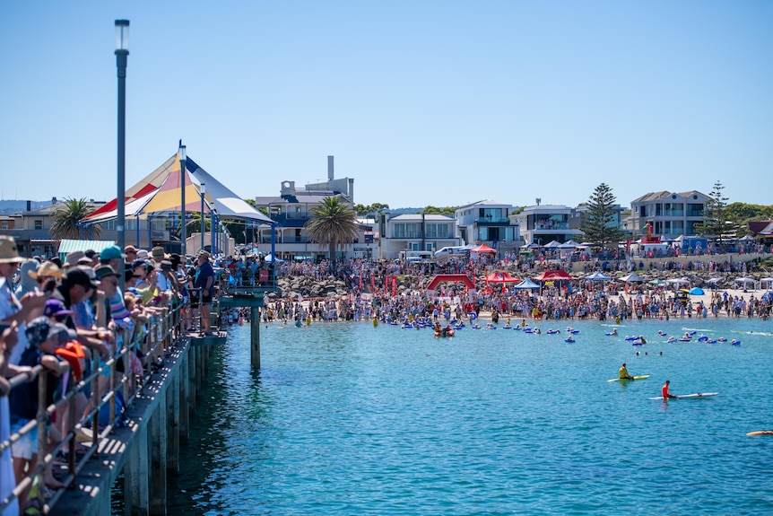 Hundreds of swimmers with blue floaties and blonde wigs prepare to swim in the ocean as onlookers crowd the Brighton jetty..