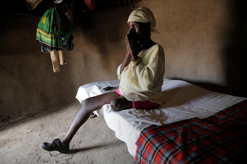 Wionnie sits on her bed in a mud floor shack with the stump of her leg visible without its prosthetic 