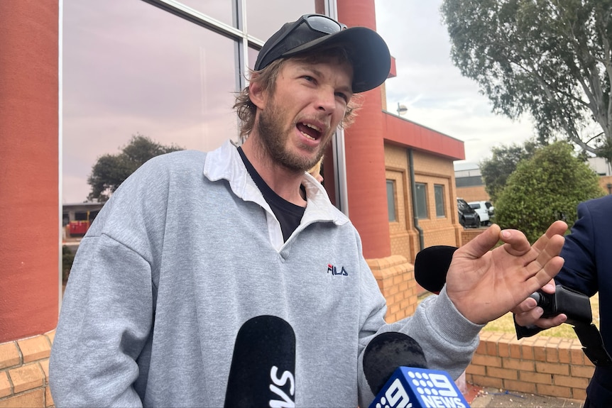 A man wearing a grey jumper and black hat addresses media