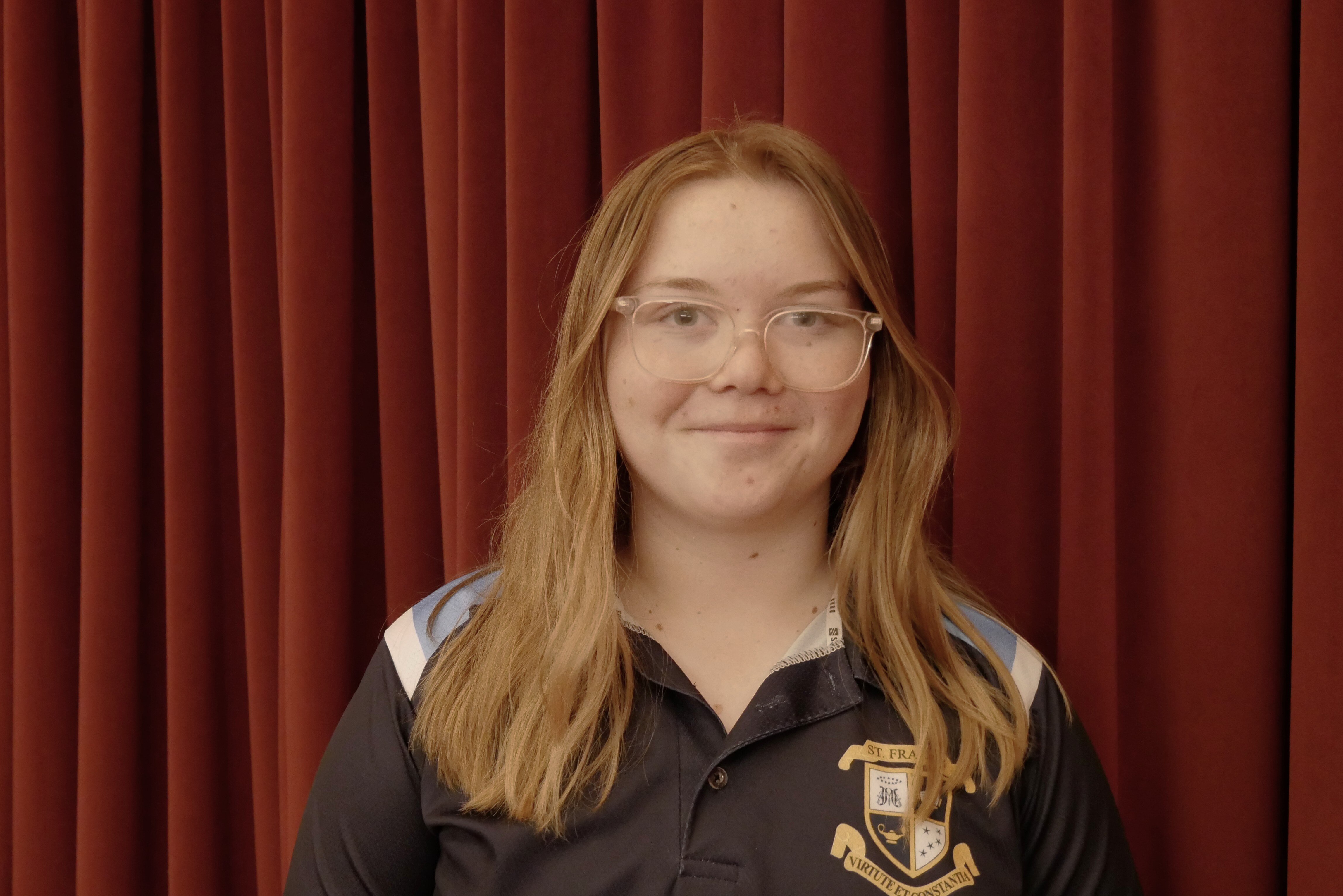 Girl in blue and black school uniform and glasses stands in front of a red curtain.