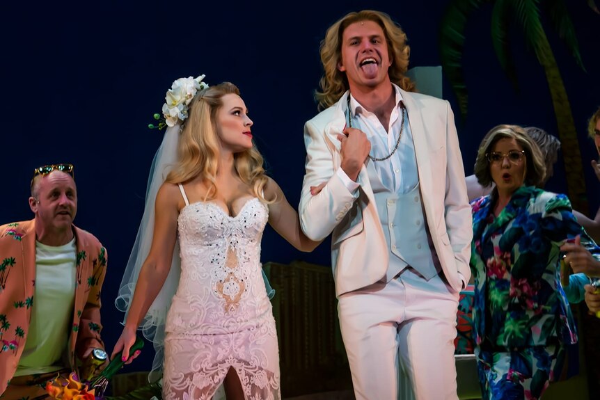 A blonde white woman in a wedding dress looks intensely at a blonde white man in a white suit, sticking his tongue out, onstage.
