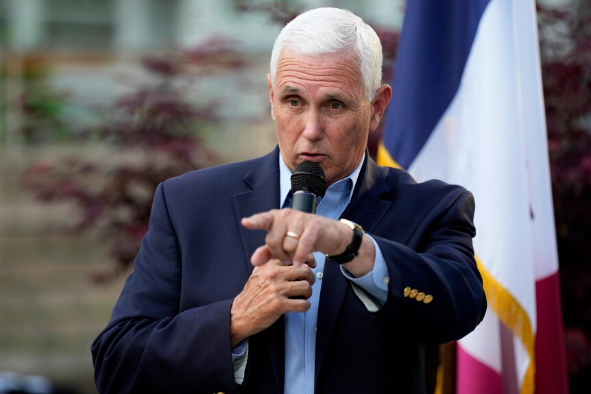 A man with short white hair wearing suit points while speaking into a microphone.