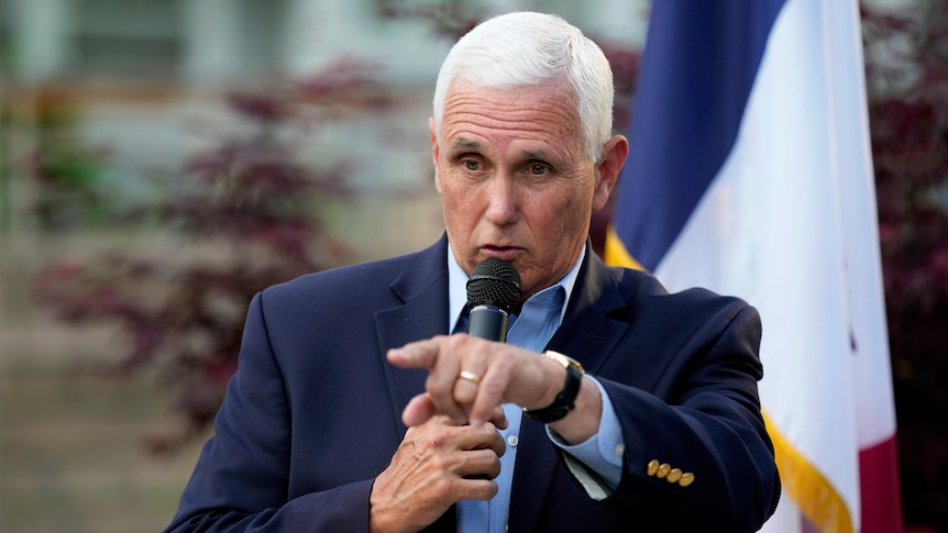 A man with short white hair wearing suit points while speaking into a microphone.