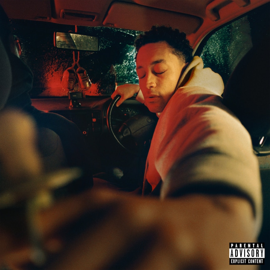 loyle carner in the drivers seat of a car, turning to the backseat with his eyes closed