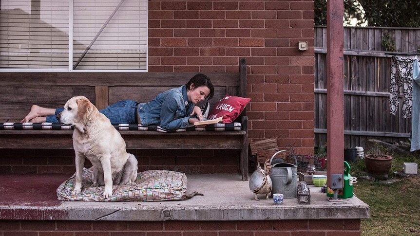 Colour photograph of person lying on a wooden bunch on balcony reading a book next to their Labrador.
