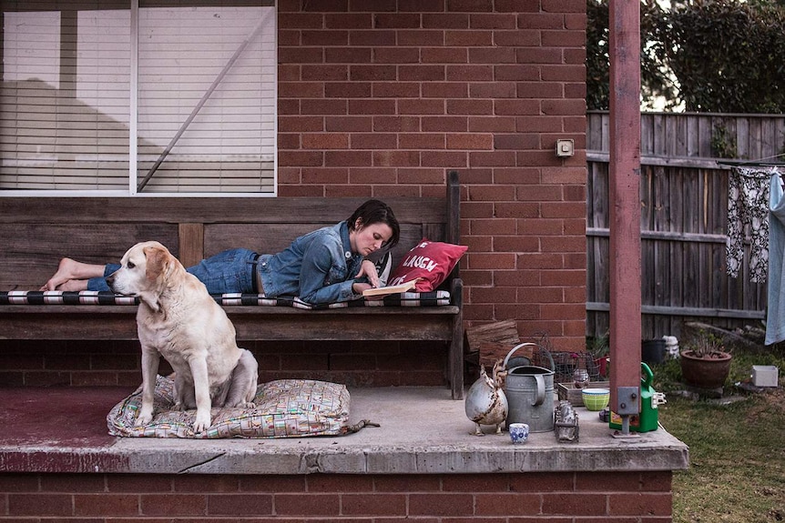 Colour photograph of person lying on a wooden bunch on balcony reading a book next to their Labrador.