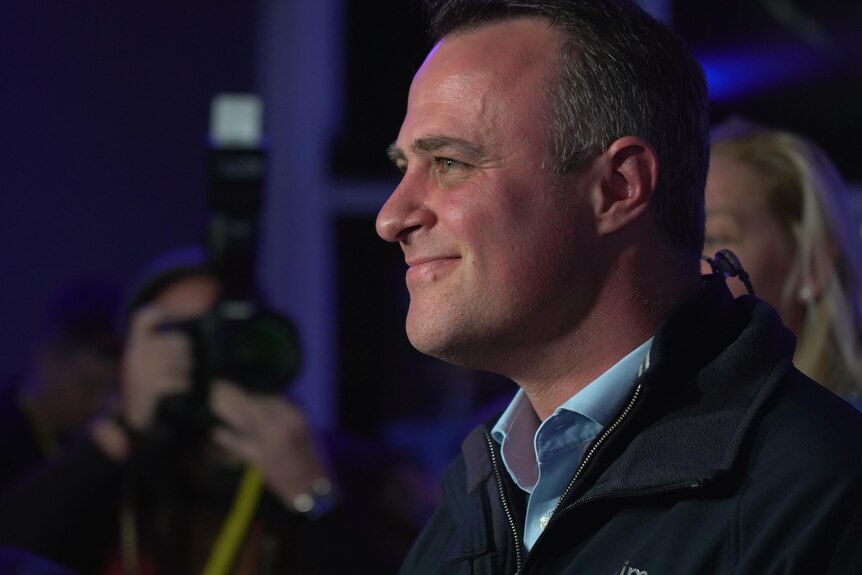 Tim Wilson smiles as he stands in front of supporters, in a blue shirt and black jacket.