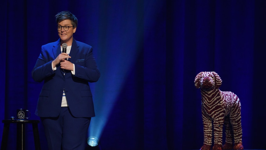 Comedian with short hair and glasses holding mic wearing blue suit, beside life-sized sculpture of dog.