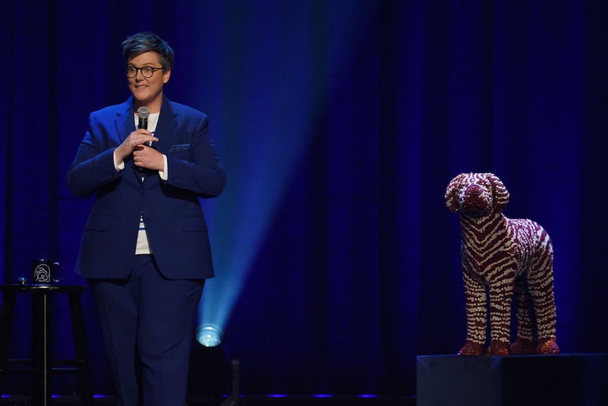 Comedian with short hair and glasses holding mic wearing blue suit, beside life-sized sculpture of dog.