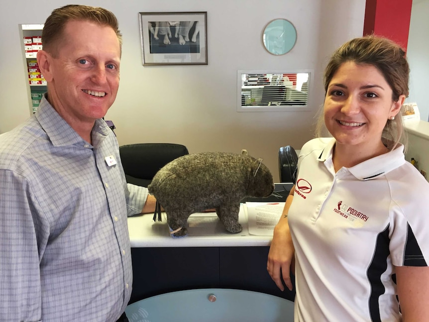 A man and a woman lean on a counter and smile at the camera. In between them, a toy wombat stands with a miniature orthotic.