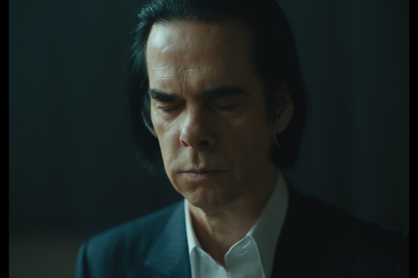 A close-up of Nick Cave, a middle-aged man in a suit, black hair pushed back from his face, with his eyes closed in reflection