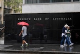 A man holding a red umbrella and two women walk past a black wall that reads Reserve Bank of Australia.
