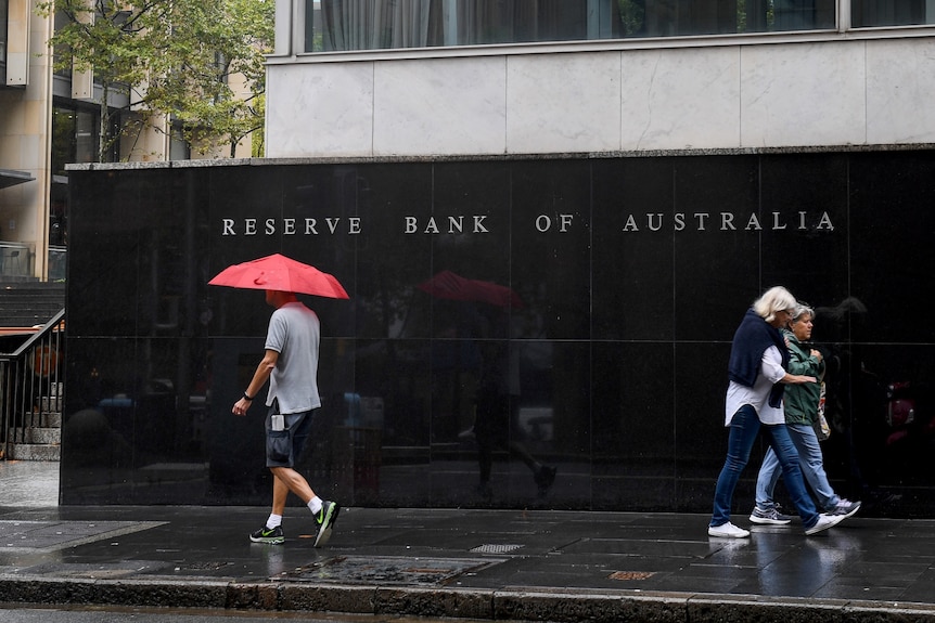 A man holding a red umbrella and two women walk past a black wall that reads Reserve Bank of Australia.