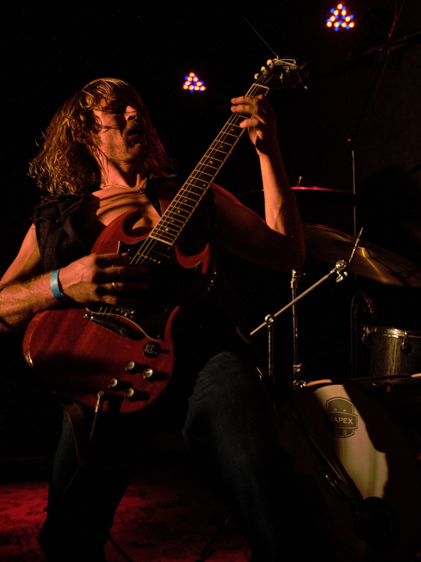 A man with long hair plays guitar on stage.