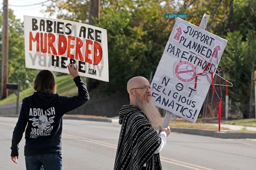 Two people, one holding an anti-abortion sign and the other holding a pro-abortion sign, stand on the street