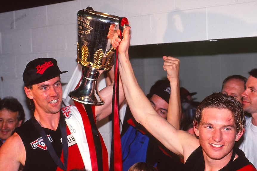 Two AFL players in the rooms after a grand final, surrounded by people as they hold up the cup.