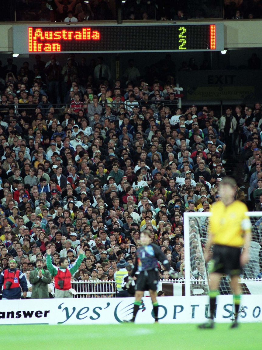 A Socceroos player stands on the pitch, with a quiet MCG crowd in the background, and a scoreboard saying 'Australia 2 Iran 2'.