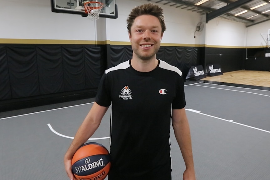 A Melbourne United NBL players stands holding a basketball to his right side.