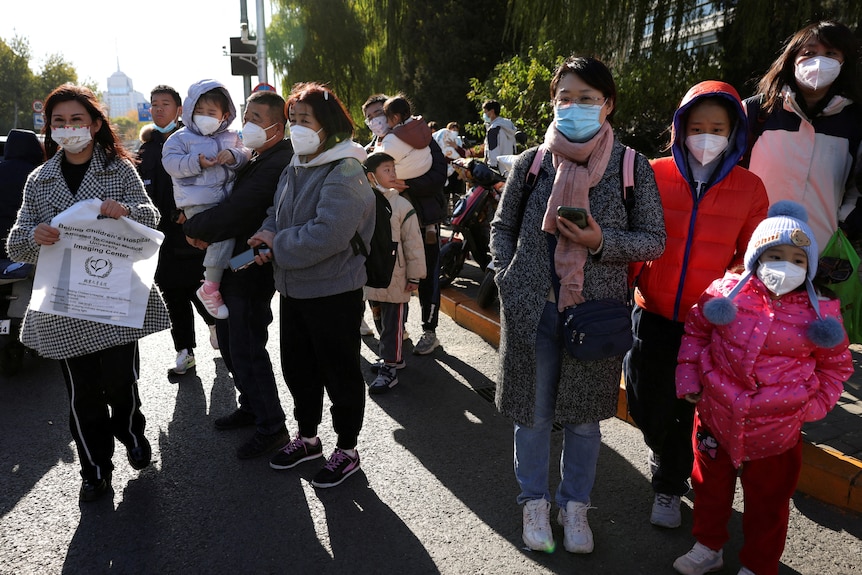 A group of adults and children wearing masks stand outside wearing jackets and coats.