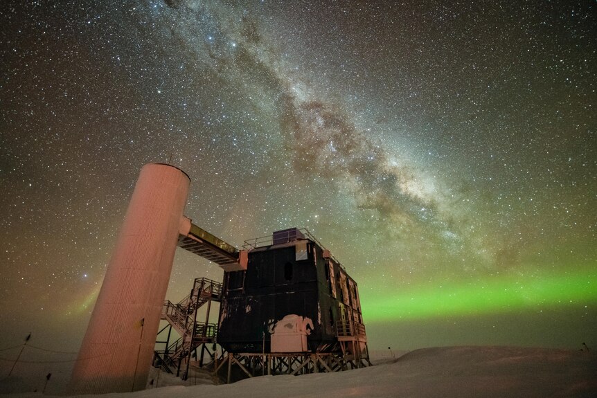 The IceCube Lab is seen under a starry, night sky, with the Milky Way appearing over low auroras in the background.