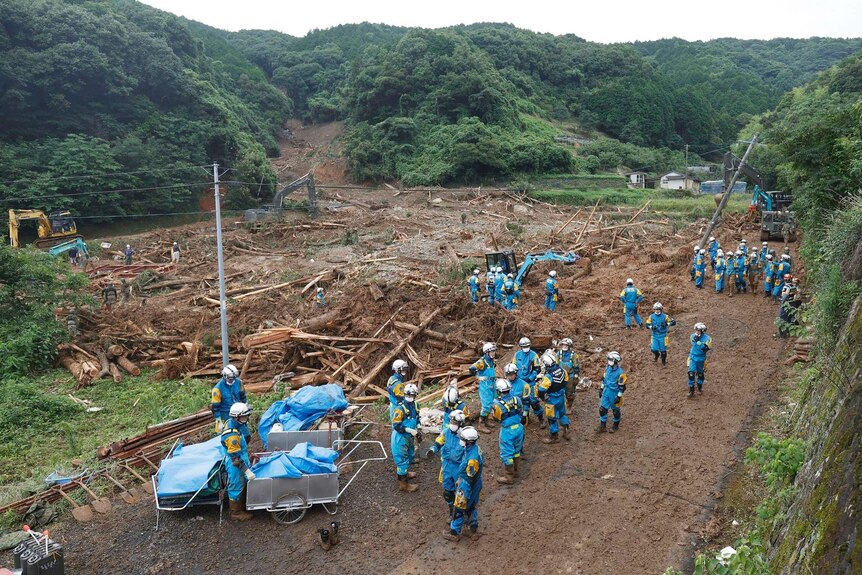 Rescue teams in blue overalls clear fallen logs and debris at the site of a mudslide.