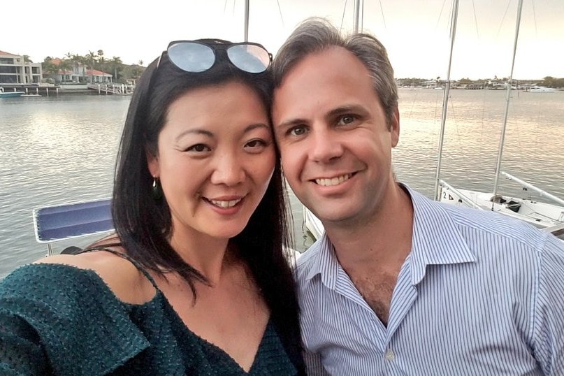 Lillian Leigh with her partner, he is a man in a business shirt and they are standing together in front of Sydney harbour.