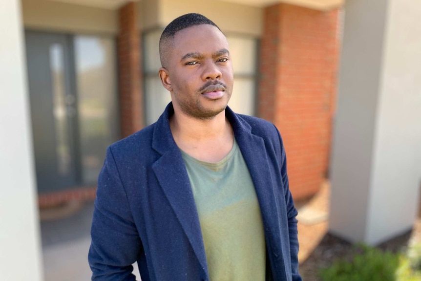 A man with short hair, wearing a jacket, stands outside a brick house.