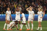 Six feamle soccer players, wearing white, look upset after losing, with one on her knees, crying