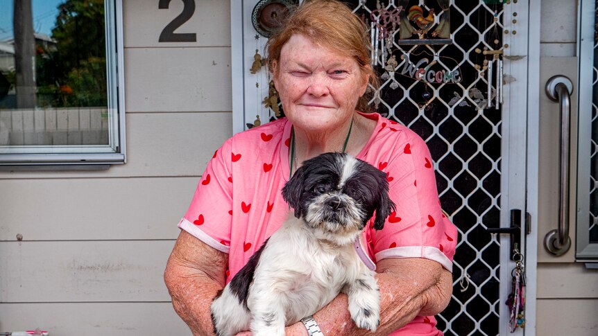 An older woman in a bright shirt sits out the front of her home, cradling a dog.