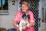 An older woman in a bright shirt sits out the front of her home, cradling a dog.